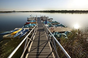 Eine Holzstege führt zu einem ruhigen See, wo Kajaks und Stand-Up-Paddle-Boards darauf aufgereiht sind.
