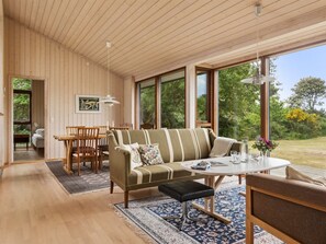 Et moderne interiør med træloft, sofa, spisebord og store vinduer med udsigt over naturen.