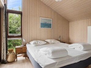 Et soveværelse med træloft, en seng med hvide sengetæpper, et natbord, en lampe og et vindue med gardiner.