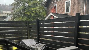 Ett trädäck med en bänk och en överbyggd sittplats, en byggnad i tegel som syns i bakgrunden och ett trästaket.