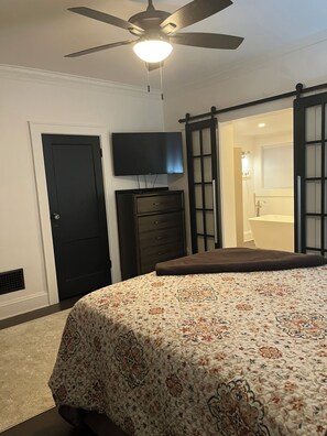 Un dormitorio con cama, televisor, cómoda, puerta y puerta corredera que da a un baño.