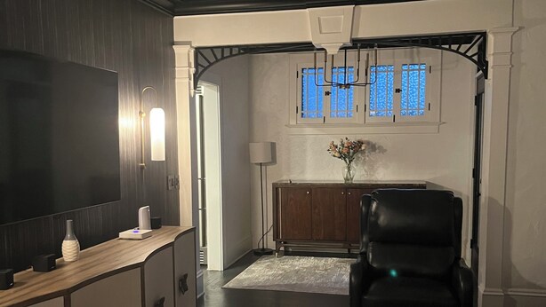 Ett vardagsrum med en platt-TV, träskåp, en svart lädersoffa, en matplats med bord och stolar samt ett fönster med blåtonat glas.