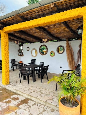 Una terrazza esterna con tetto di paglia, un tavolo con sedie, specchi decorativi e piante in vaso.