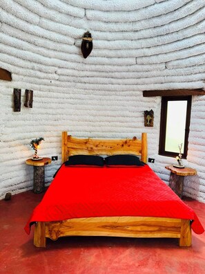 Una stanza con un letto di legno, pareti bianche dalla trama decorativa, una finestra e due piccoli comodini in legno con lampade.