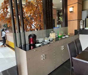 A buffet setup with a coffee maker, plates, and a green container on a counter. - Cozzy Menara Archie (Ternate)