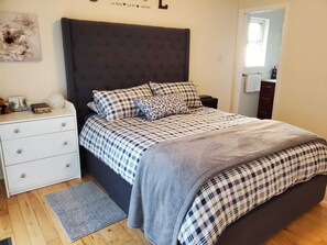 Ein Schlafzimmer mit dunklem Kopfteil, einem Bett mit karierter Bettwäsche, einem Nachttisch mit Lampe sowie einem kleinen Teppich.