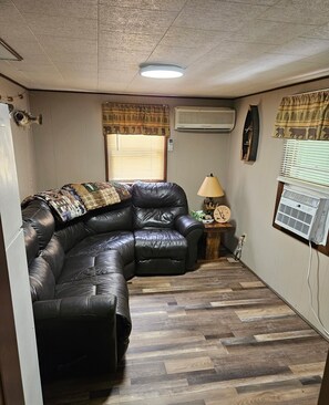 A living room with a black leather sectional sofa, a wooden floor, an air conditioning unit, and a lamp on a side table.