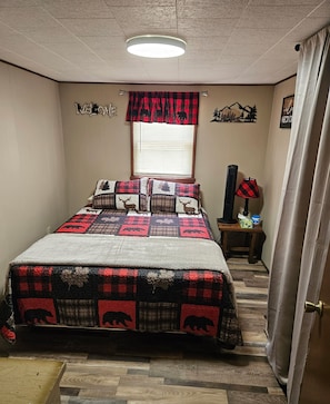 Ein Schlafzimmer mit einem Bett, einem Fenster mit Jalousien, einer Wanduhr, einer Lampe und einem kleinen Tisch mit einem dekorativen Gegenstand.