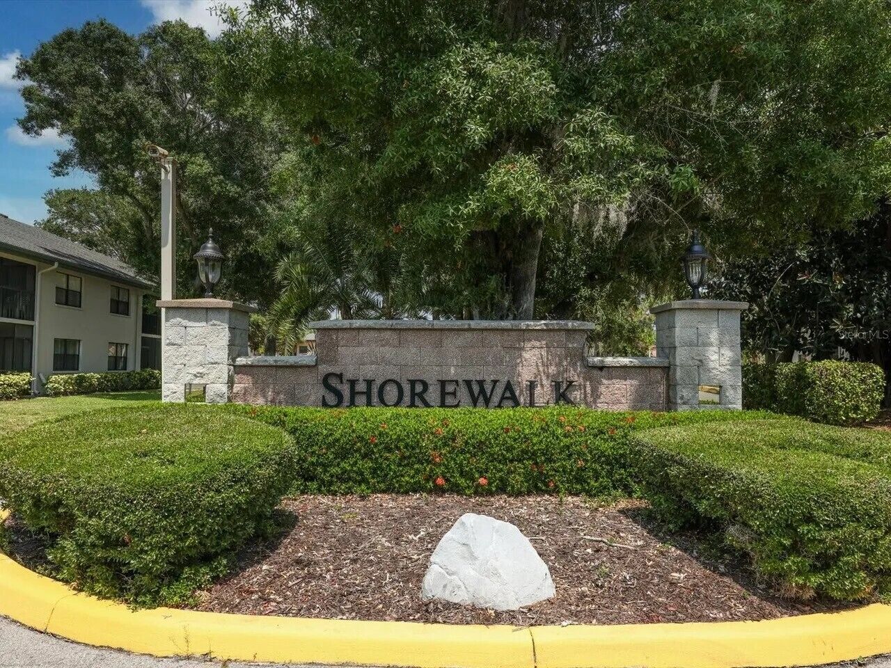 Une pancarte indiquant « SHOREWALK » est placée devant un bâtiment, entouré de verdure et bordé d’un trottoir jaune.