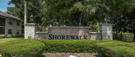 Une pancarte indiquant « SHOREWALK » est placée devant un bâtiment, entouré de verdure et bordé d’un trottoir jaune.
