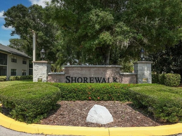 A sign with 'SHOREWALK' in front of a building, surrounded by greenery and a yellow curb.