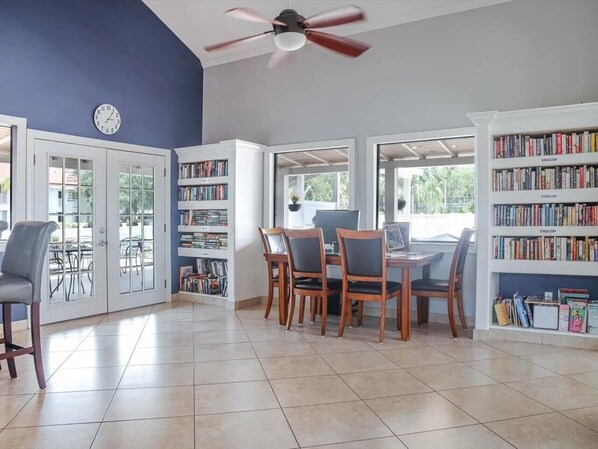 A room with a dining table, chairs, a ceiling fan, a bookshelf, and a clock on the wall.