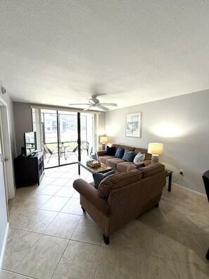 A living room with a brown sofa, a coffee table, a ceiling fan, and a view through a glass door to an outdoor patio.