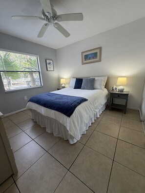 A bedroom with a bed, two bedside tables, a ceiling fan, a window with blinds, and a painting on the wall.