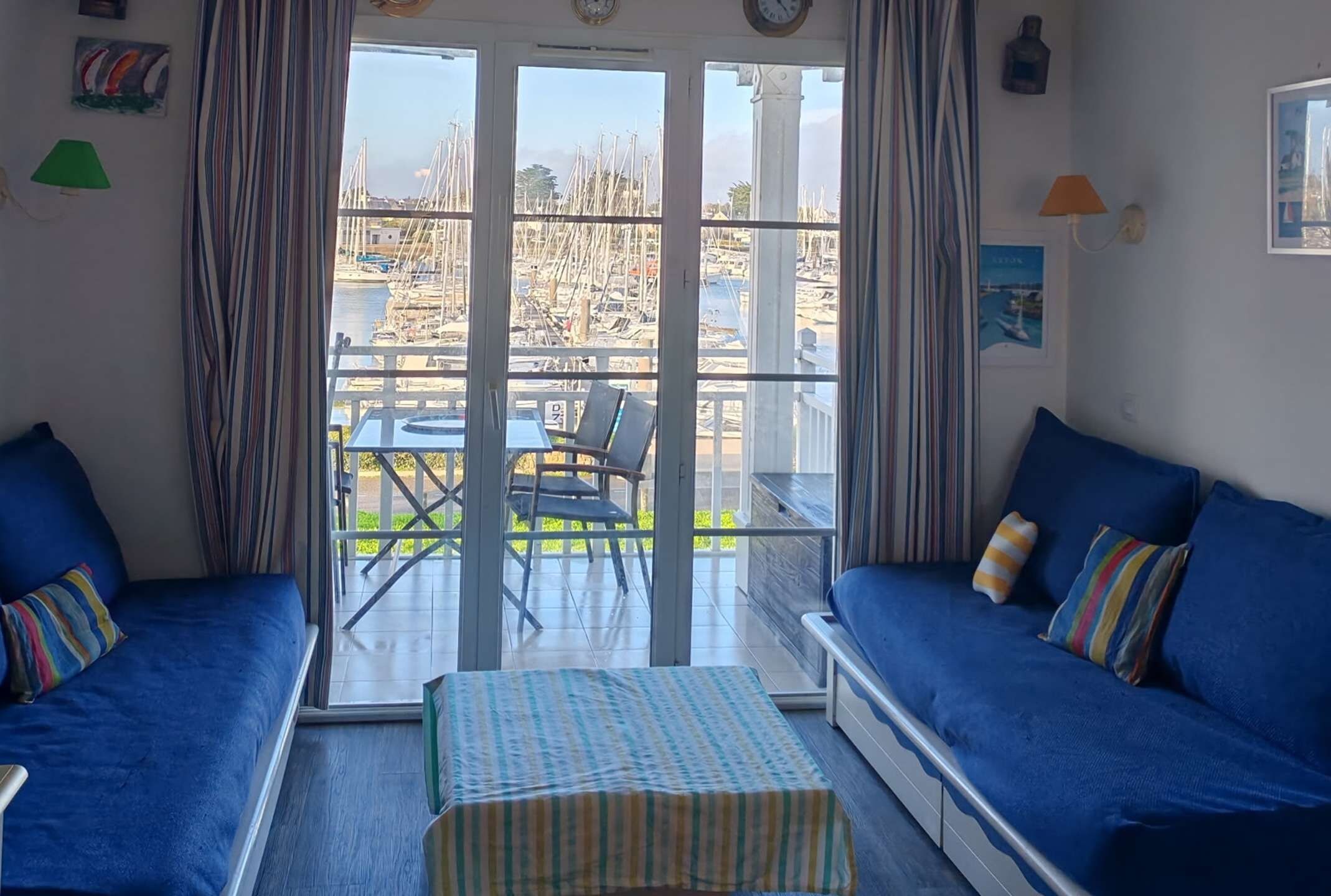 A room with a sliding glass door leading to an outdoor area with a view of boats and a dock. There are two blue sofas with striped cushions and a striped rug on the floor.