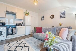 Et moderne køkken med mikrobølgeovn, ovn og integreret vaskemaskine. Der er en grå sofa med puder og en dekorativ vase med blomster.