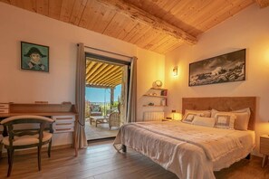 Ein Schlafzimmer mit Holzbalkendecke, einem Bett mit Kissen, einem Schreibtisch mit Stuhl und Blick durch eine geöffnete Tür.