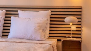 Cama bem arrumada com roupa de cama branca, cabeceira de madeira com ripas horizontais e mesa de cabeceira com abajur.