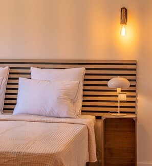 Ein ordentlich bezogenes Bett mit weißer Bettwäsche, ein Holzkopfbrett mit horizontalen Lamellen und ein Nachttisch mit Lampe.