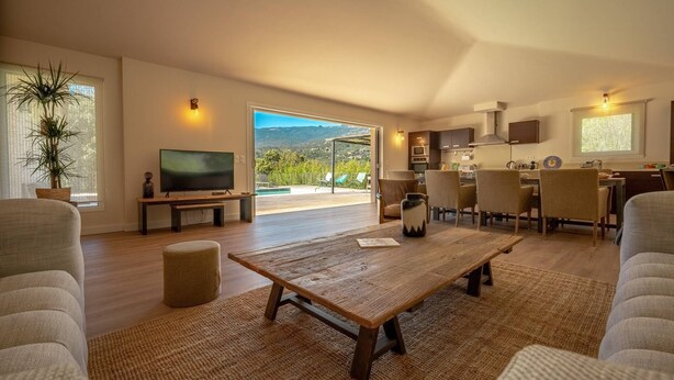 Uma sala de estar ampla com uma TV grande, assentos confortáveis, uma mesa de centro de madeira e vista para o exterior.