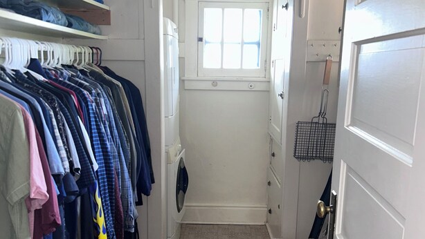 Een smalle gang met een houten vloer, een witte deur en een raam. Kleding hangt op een rek, en er staat een schoenenrek.