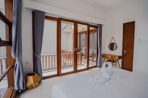 A hotel room with a bed, a wooden desk, a small table, a balcony with a railing, and a sliding glass door.