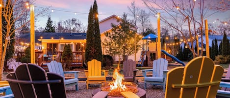 An outdoor seating area with Adirondack chairs, a fire pit, and string lights against a twilight sky.