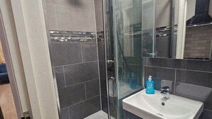 A modern bathroom with a glass shower enclosure, a sink with a mirror, and dark-toned tiles. - Stylish Brick Lane Studio Near Tube & Dining Hub! (London)