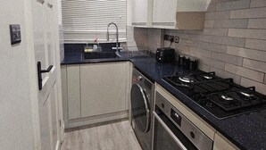 A compact kitchen with a stove, oven, and sink, featuring a tiled backsplash and a window with blinds. - Stylish Brick Lane Studio Near Tube & Dining Hub! (London)