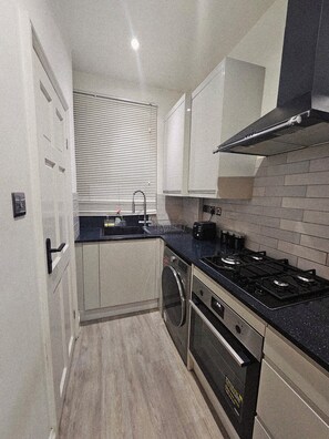 A compact kitchen with a stove, oven, and sink, featuring a tiled backsplash and a window with blinds. - Stylish Brick Lane Studio Near Tube & Dining Hub! (London)