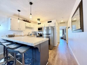 A modern kitchen with white cabinets, a stainless steel refrigerator, a large island with bar stools, pendant lighting, and a hallway leading to another room. - Las Palmas Resort #1412 | All-Inclusive Amenities (Saint George)