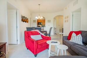 A living room with a red sofa, a grey sofa, and a small white table. There is a kitchen in the background with wooden cabinets and a microwave. - Modern 3BR VistaCay Apartment Near Disney, Universal & Orlando Attractions Sleeps 7 (Orlando)