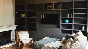Ett vardagsrum med en öppen spis, ett soffbord i trä, en soffa med mönstrade kuddar, en tv på väggen och en dekorativ taklampa.