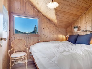 A wooden cabin room with a bed, a wicker chair, a window with a view, and a shelf with a plant. - "Greta" - 300m from the sea by Interhome (Thisted)