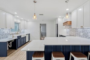 Et moderne køkken med hvide skabe, en stor ø, barstole, hængende lamper og et mønstret backsplash af fliser.