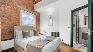 Ein Schlafzimmer mit Backsteinwand, einem großen Bett, einem Fenster, einem Heizkörper und einer Tür, die ins Badezimmer führt.