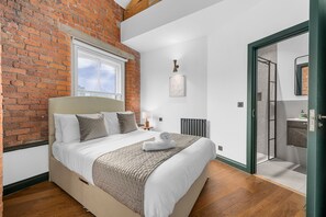 A bedroom with a brick wall, a large bed, a window, a radiator, and a door leading to a bathroom. - Spacious 2BR • Pool Table • Workspace • City Centre (Manchester)