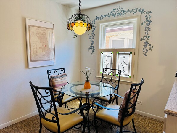 A dining area with a glass table, metal chairs, a hanging light fixture, a framed map, and a window with decorative vines.