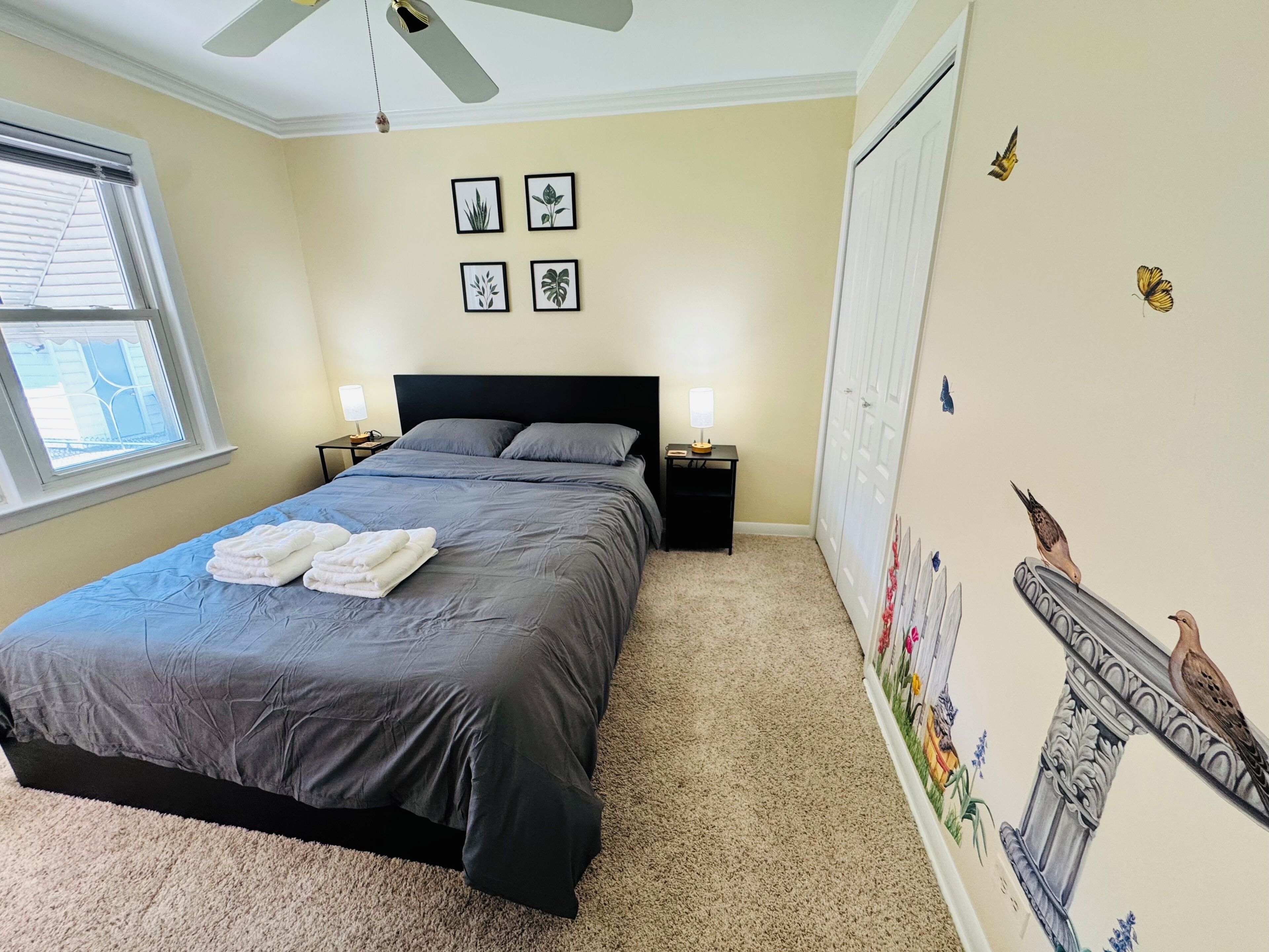 A bedroom with a bed, two bedside tables, a ceiling fan, a window, and wall decorations.