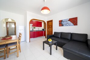 Ein modernes Wohnzimmer mit einer schwarzen Couch, einem Holzespeisesaal mit Stühlen, einem kleinen Couchtisch mit Früchten, einer Küche mit roten Schränken und einer roten Pendelleuchte.