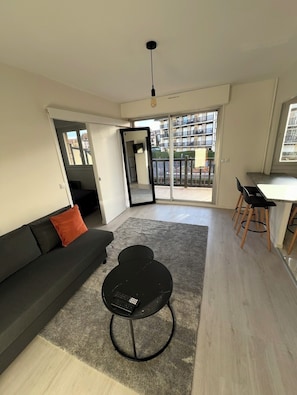 Ein modernes Wohnzimmer mit einer Couch, einem kleinen runden Tisch, einer Essecke mit Hocker und einer Glastür zum Balkon.