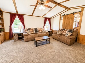 A cozy living room with a sofa set, a coffee table, wooden beams, a ceiling fan, and a view through a window.