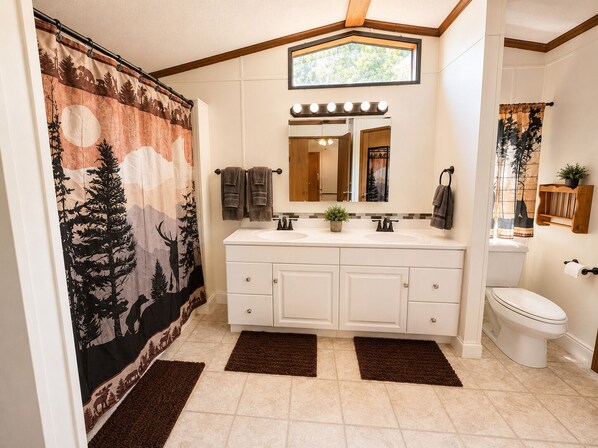A bathroom with a double sink vanity, a shower curtain with a forest design, a mirror with lights, and a view of the outdoors through a window.