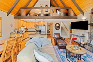 A spacious living area with a high ceiling, wooden beams, a sofa, a coffee table, a fireplace, and a kitchen in the background. - A-Frame w/ AC | Walk to Glaze Pool | Bunk Room (Sisters)