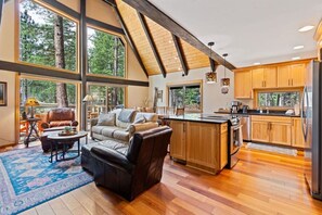 A spacious living area with a fireplace, leather sofa, and a large window overlooking a forest. - A-Frame w/ AC | Walk to Glaze Pool | Bunk Room (Sisters)
