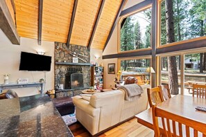 A cozy living room with a stone fireplace, a flat-screen TV, a dining area with a large window overlooking a forest. - A-Frame w/ AC | Walk to Glaze Pool | Bunk Room (Sisters)