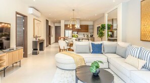 Ein modernes Wohnzimmer mit Sofa, Couchtisch und Fernseher. Ein Essbereich mit Stühlen und Tisch ist ebenfalls vorhanden. Im Hintergrund ist die Küche sichtbar.