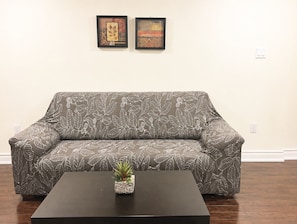 En sofa med bladmotiv, en lille plante på et bord og to billeder i rammer på væggen.