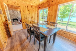 A wooden cabin interior with a dining table, chairs, a fireplace, and a large window overlooking a lake.
