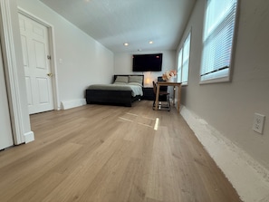 Ein Schlafzimmer mit einem Bett, einem Schreibtisch, einem Stuhl, einer Tür und einem Fenster mit Jalousien.